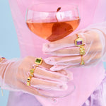 Person holding a glass of pink drink with transparent gloves and colorful jewelry against a light blue background