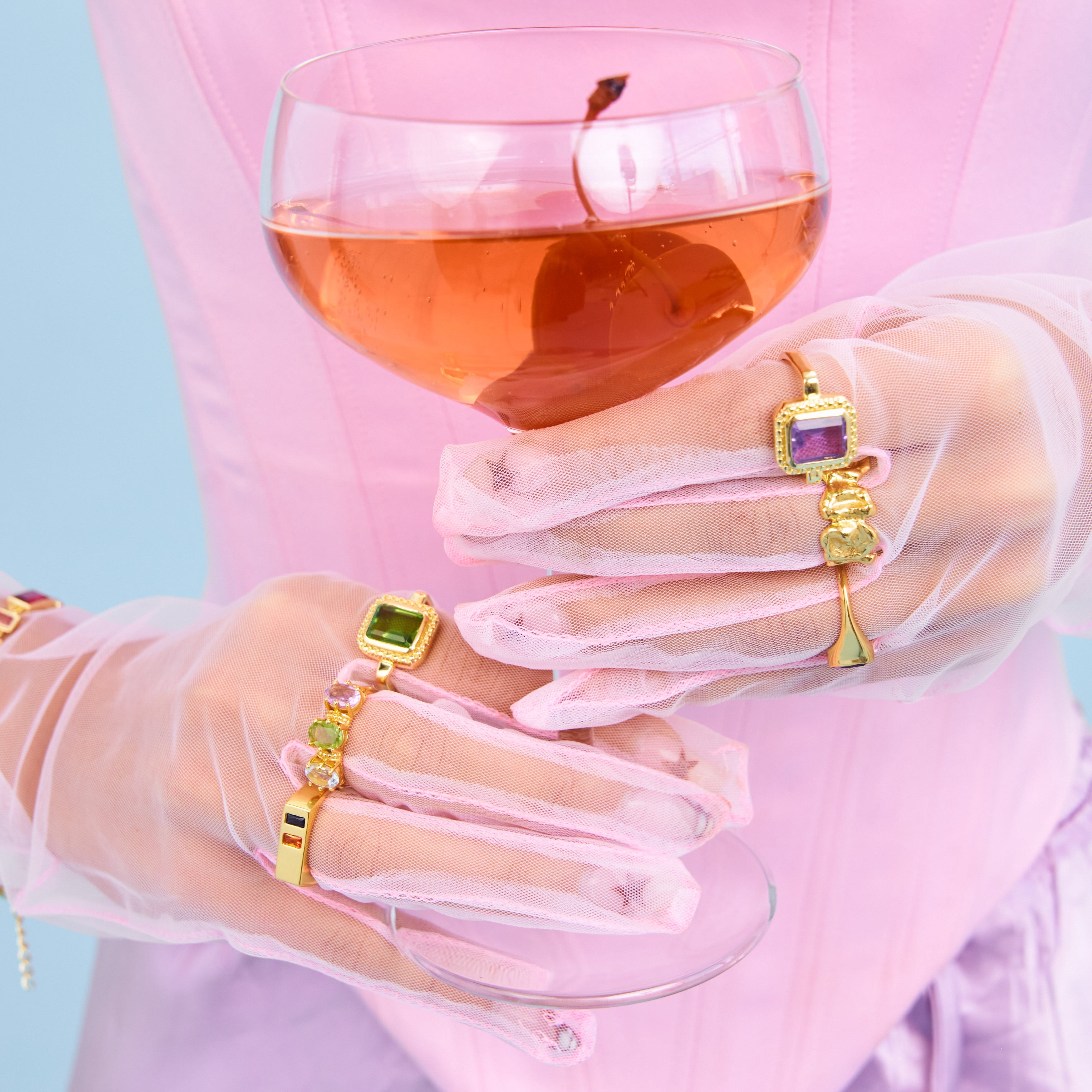 Person holding a glass of pink drink with transparent gloves and colorful jewelry against a light blue background