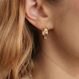 A close-up of a model's ear wearing a small gold hoop earring with a pink gemstone inset.