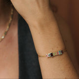 A close-up lifestyle shot of a woman's hand and wrist wearing a delicate gold bracelet with a colorful rectangular stone accent, alongside matching gold hoop earrings.
