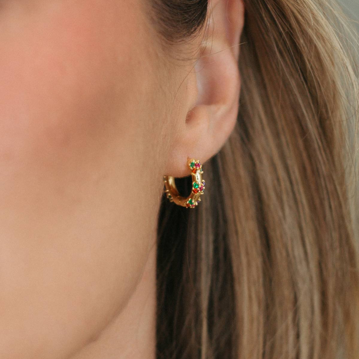 A close-up of a woman's ear wearing a small gold crescent-shaped hoop earring with a black center and tiny red and orange stones.