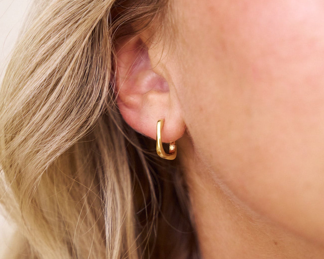 Close-up of a person wearing a square gold hoop earring with blonde hair.