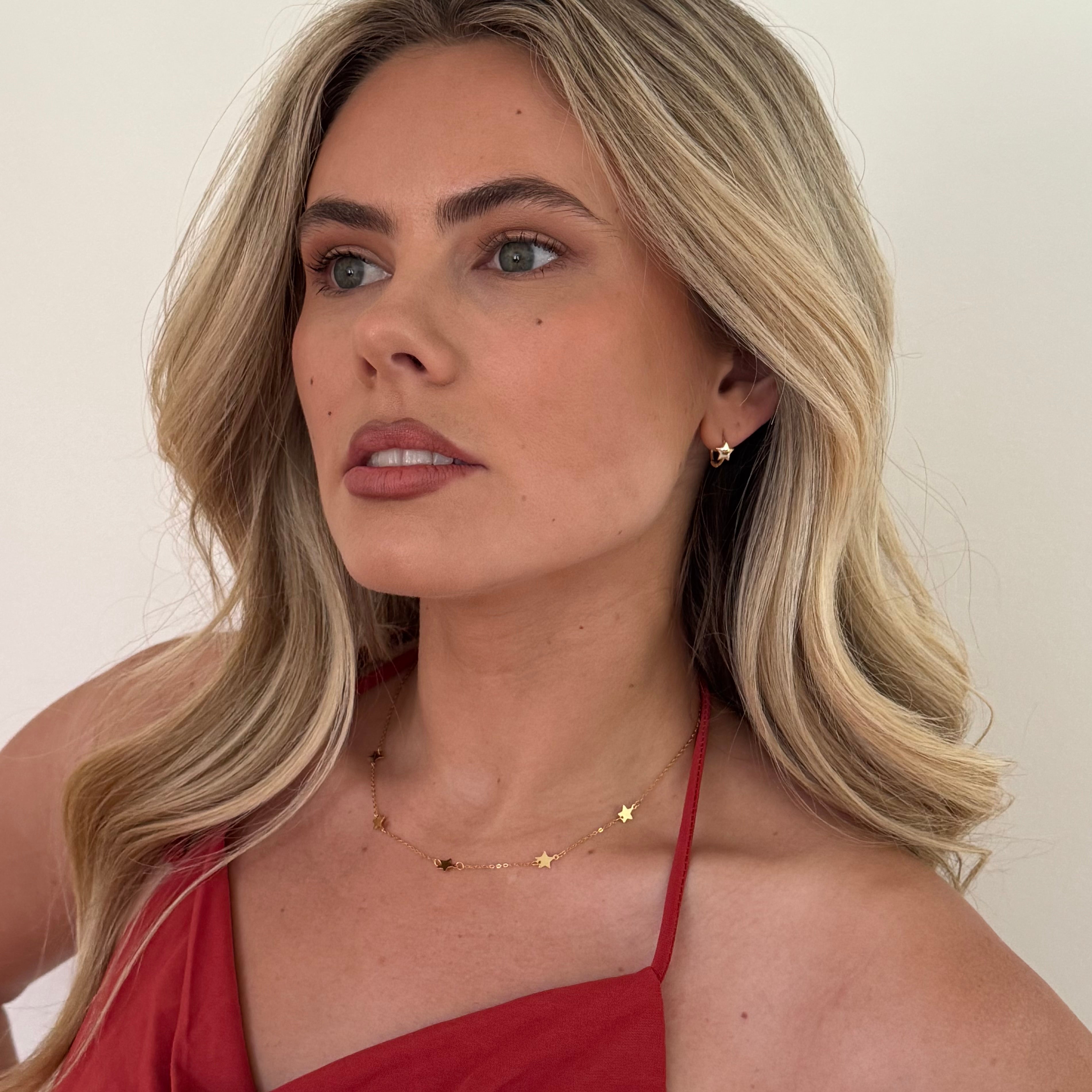 Woman with blonde hair wearing a red top and gold jewelry against a plain background