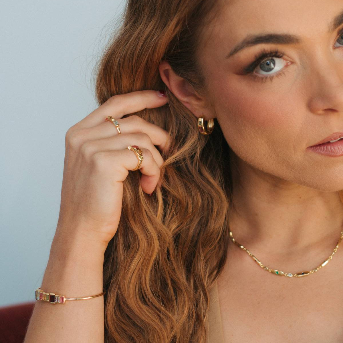 A close-up lifestyle shot of a woman with long wavy light brown hair, wearing a delicate gold chain necklace, a gold huggie hoop earring, and a gold ring on one finger. She is wearing a strapless top against a soft, light blue-gray background.