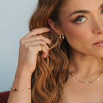 A close-up lifestyle shot of a woman with long wavy light brown hair, wearing a delicate gold chain necklace, a gold huggie hoop earring, and a gold ring on one finger. She is wearing a strapless top against a soft, light blue-gray background.