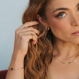 A close-up lifestyle shot of a woman with long wavy light brown hair, wearing a delicate gold chain necklace, a gold huggie hoop earring, and a gold ring on one finger. She is wearing a strapless top against a soft, light blue-gray background.