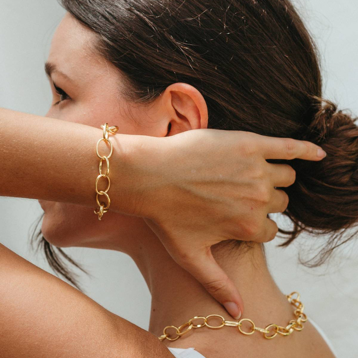 Woman wearing gold chain bracelets on her wrist and neck against a neutral background