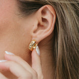 A close-up of a woman's ear wearing a small gold huggie hoop earring, with her hand near her face.