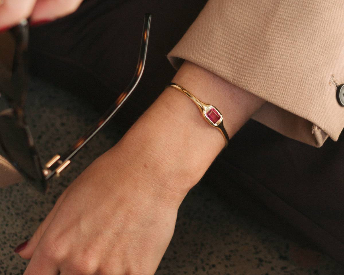 A close-up lifestyle shot of a person wearing a gold bangle bracelet with a rectangular red gemstone, paired with a beige blazer and black trousers.