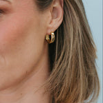 A close-up of a woman wearing a gold hoop earring, with her ear, hair, and neck visible against a light blue background.