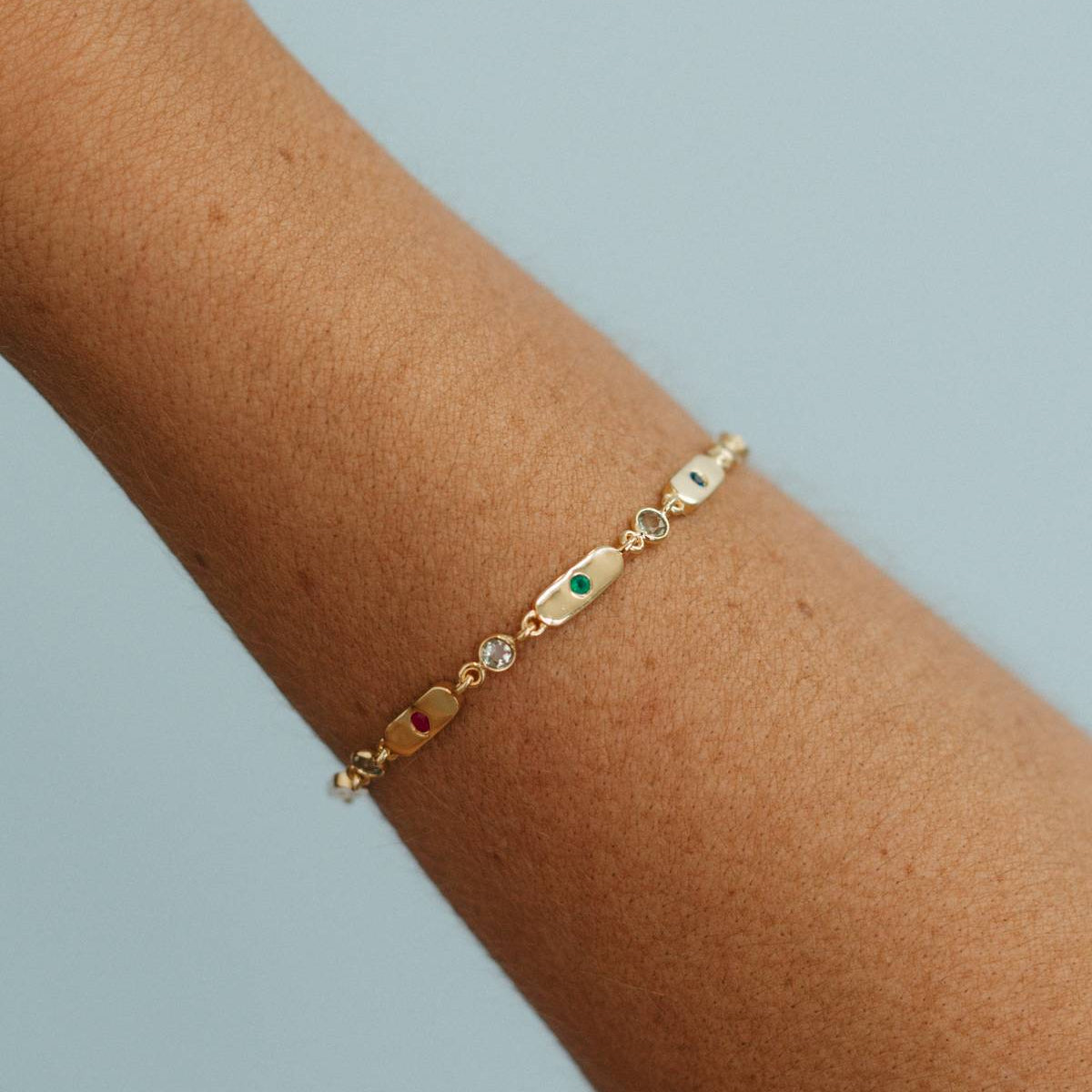 A delicate gold chain bracelet with small oval charms, including a green stone and a clear crystal, worn on a person's wrist against a light blue background.