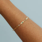 A delicate gold chain bracelet with small oval charms, including a green stone and a clear crystal, worn on a person's wrist against a light blue background.