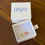 A gold ring with a pink heart-shaped gemstone and a small green stone, displayed inside an open white jewelry box with the brand name 'BIXBY' printed on the lid, set on a wooden table.