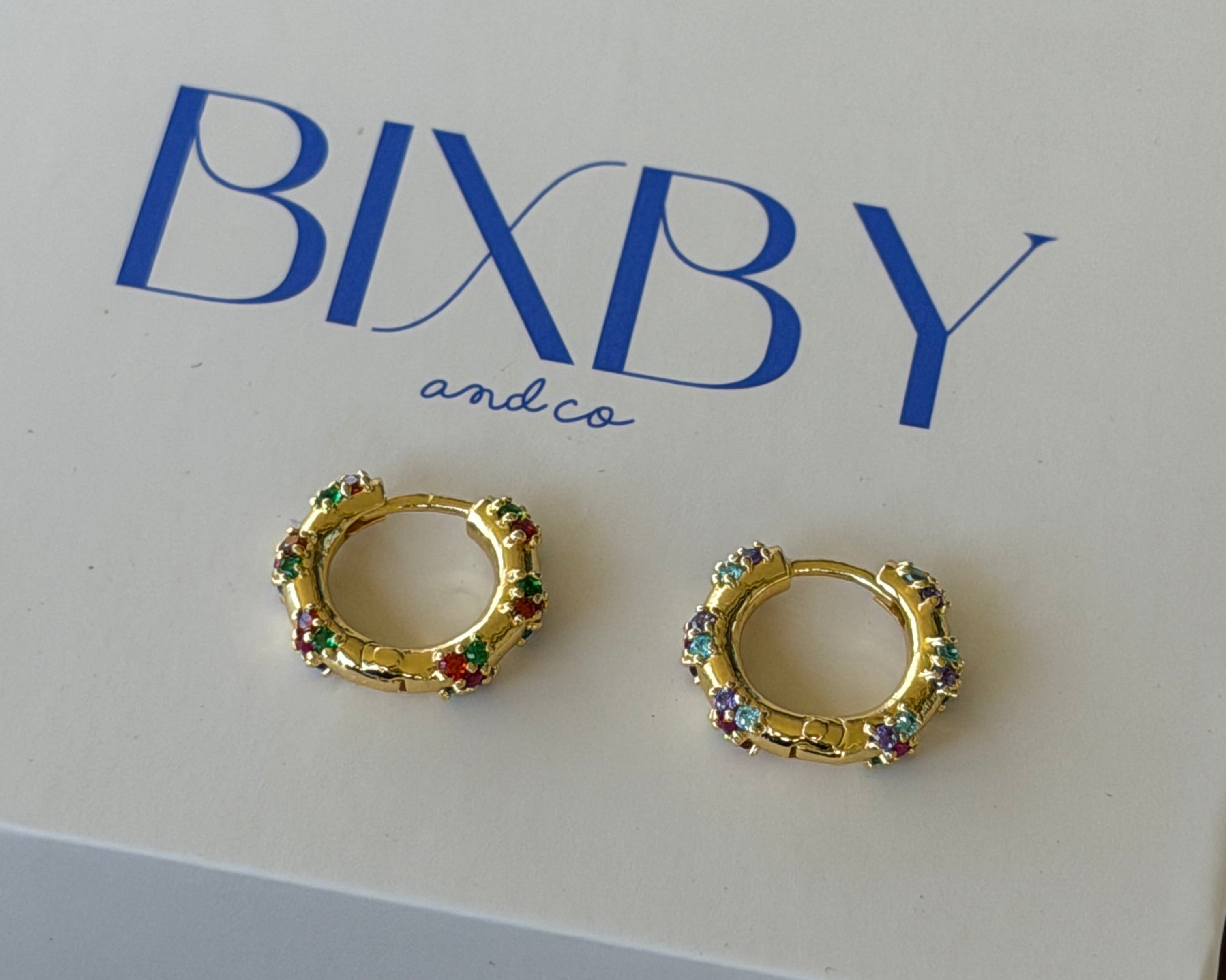 A pair of gold huggie hoop earrings with colorful rhinestone accents, displayed on a white BIXBY and Co. brand gift box on a dark wooden surface.