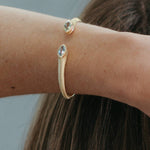 A close-up of a person's wrist wearing a gold snake-style cuff bracelet with two oval clear gemstone accents, against a soft gray background and brown hair.