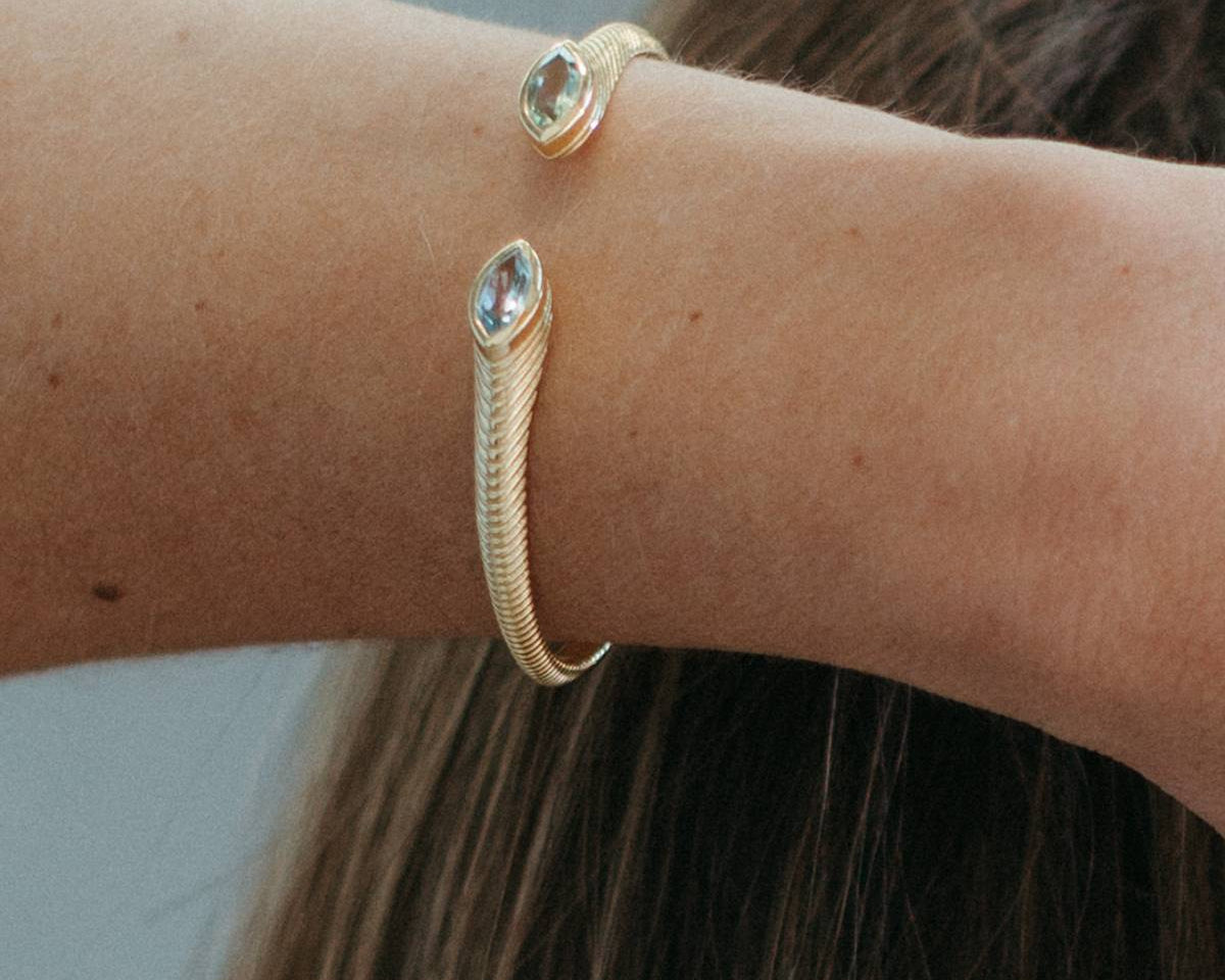 A close-up of a person's wrist wearing a gold snake-style cuff bracelet with two oval clear gemstone accents, against a soft gray background and brown hair.