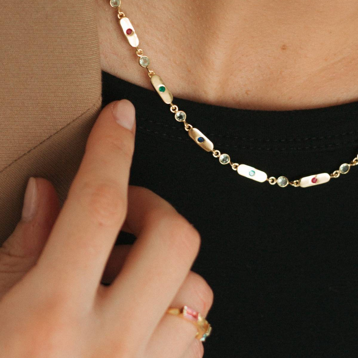 A close-up of a delicate gold chain necklace with oval mother-of-pearl links, small green and red gemstone accents, worn on a model's neck with a hand resting near the chest.
