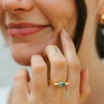 Close-up of a woman's hand wearing a gold ring with a green gemstone, touching her face.