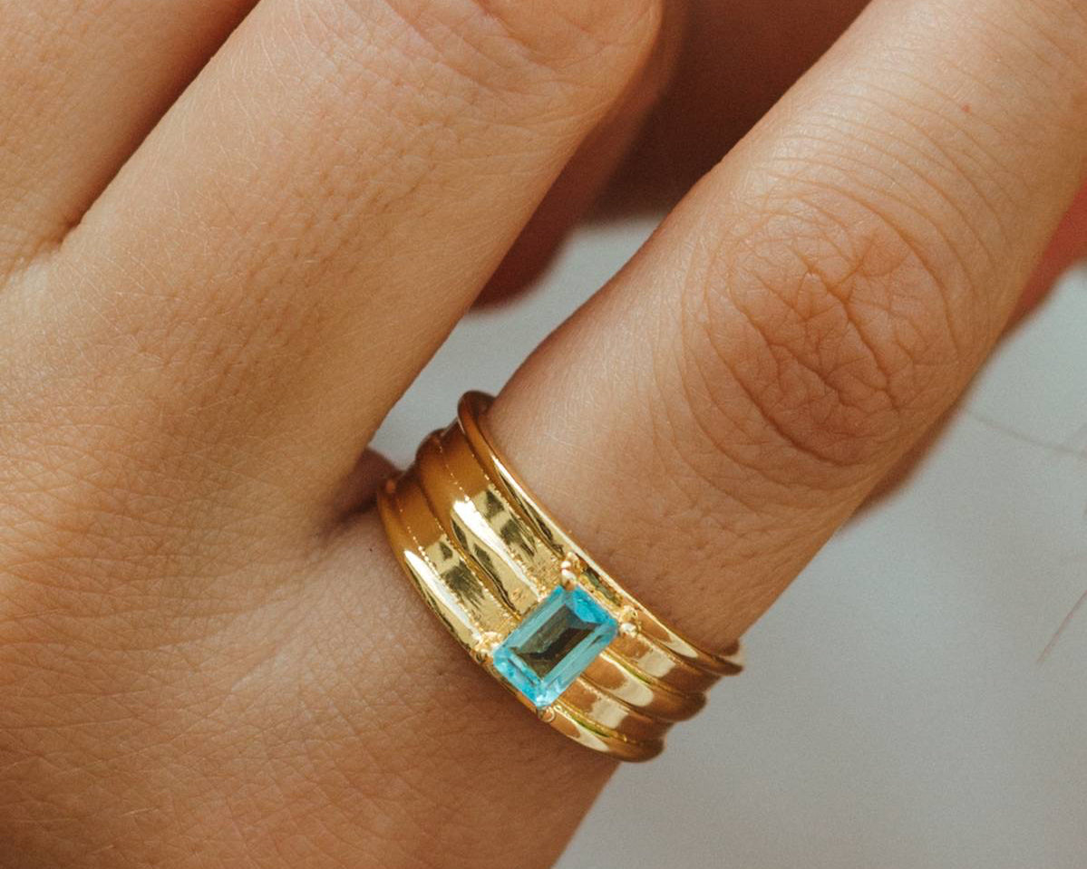 Gold ring with a blue gemstone on a person's finger against a neutral background