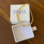A gold chain bracelet with textured, twisted links draped over an open white jewelry box, with the brand name 'bixby printed on the lid, set on a wooden table.