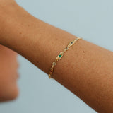 A delicate gold chain bracelet with small green stone accents, worn on a person's wrist against a soft light blue background.