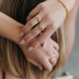 A close-up lifestyle shot of a person's hands and hair, showcasing several gold rings with small red gemstone accents, a gold beaded bracelet, and light purple nail polish against a soft gray background.
