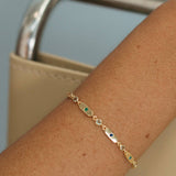 A delicate gold chain bracelet with small green and blue stones, worn on a person's wrist next to a beige handbag.