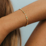 A delicate gold chain bracelet with small green and white stone accents worn on a woman's wrist, shown against a soft light blue background.