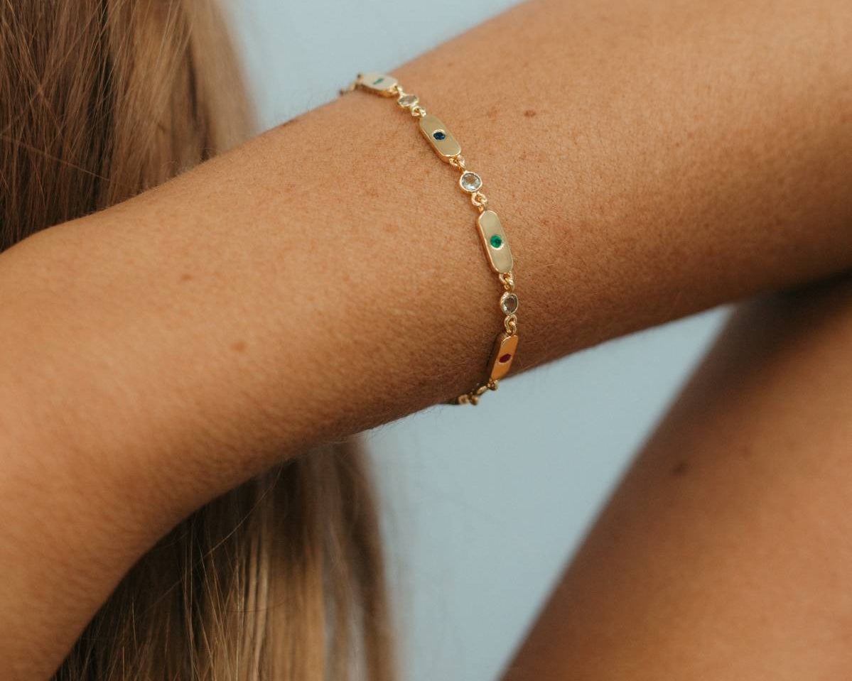 A delicate gold chain bracelet with small green and white stone accents worn on a woman's wrist, shown against a soft light blue background.