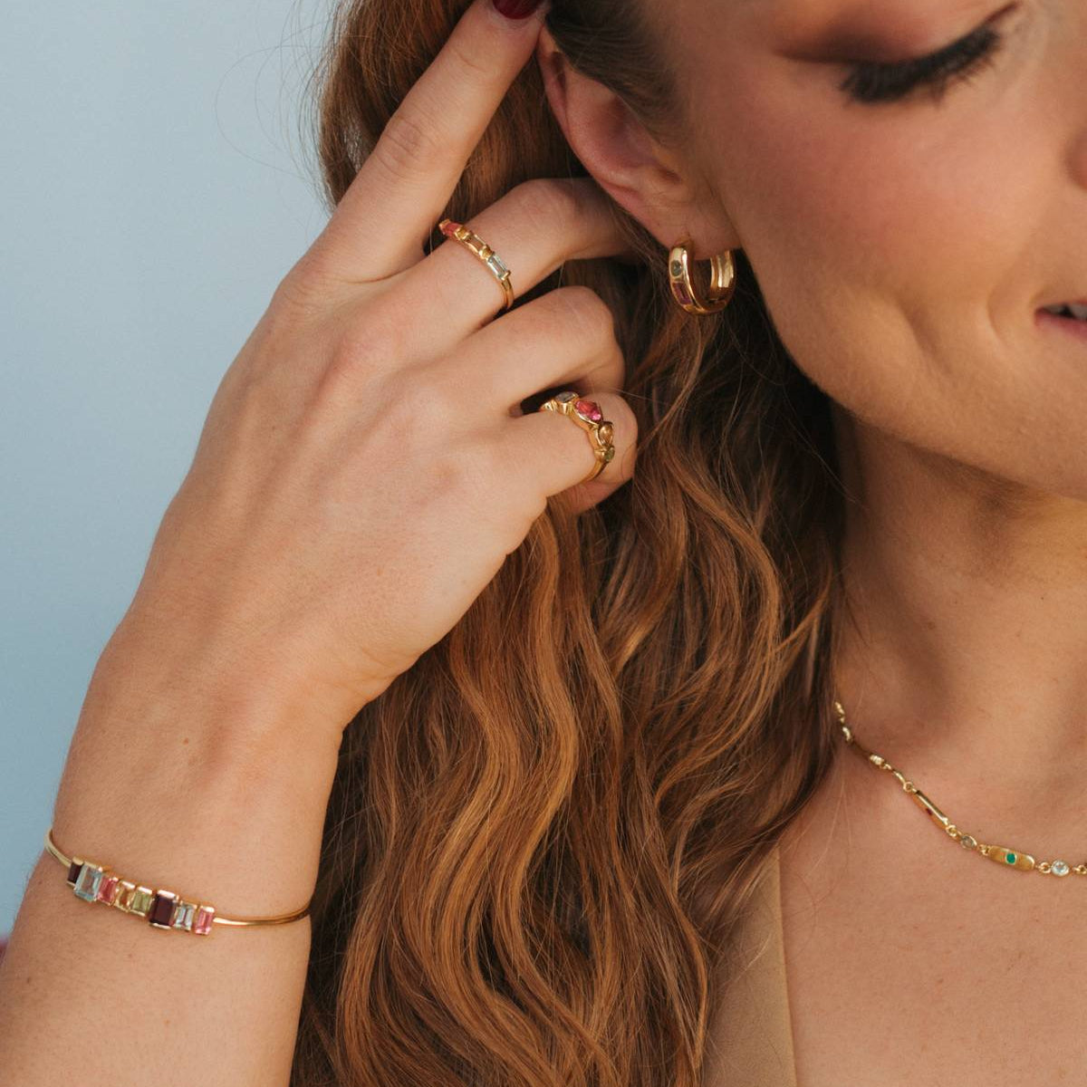 A close-up lifestyle shot of a woman with long wavy light brown hair, wearing a gold bracelet with colorful rectangular gemstones on her wrist, gold rings on her fingers, and a delicate gold necklace against a soft light blue background.