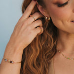 A close-up lifestyle shot of a woman with long wavy light brown hair, wearing a gold bracelet with colorful rectangular gemstones on her wrist, gold rings on her fingers, and a delicate gold necklace against a soft light blue background.