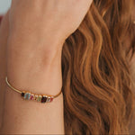 A close-up lifestyle shot of a woman wearing a delicate gold chain bracelet on her wrist, adorned with small colorful gemstone charms. She also wears gold rings and hoop earrings, with her hand resting near her ear against a soft, out-of-focus background.