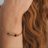 A close-up lifestyle shot of a woman wearing a delicate gold chain bracelet on her wrist, adorned with small colorful gemstone charms. She also wears gold rings and hoop earrings, with her hand resting near her ear against a soft, out-of-focus background.