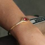 A gold-toned bangle bracelet with a red faceted gemstone, worn on a person's wrist.