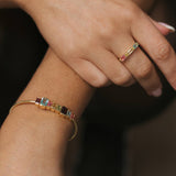 A close-up lifestyle shot of a person's hands and arms, showcasing a delicate gold bangle bracelet and matching gold ring, both embellished with small colorful gemstone accents, against a dark blurred background.
