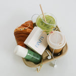 A styled flat-lay of various items arranged in a brown paper tray on a white background, including a green drink with a straw, a takeaway coffee cup, a green bottle, a white box with blue 'FANBY' branding, a croissant, and gold jewelry.