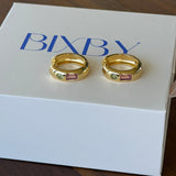 Two gold-toned BIXBY rings with small pink and green gemstones, displayed on a white BIXBY gift box on a dark wooden surface.