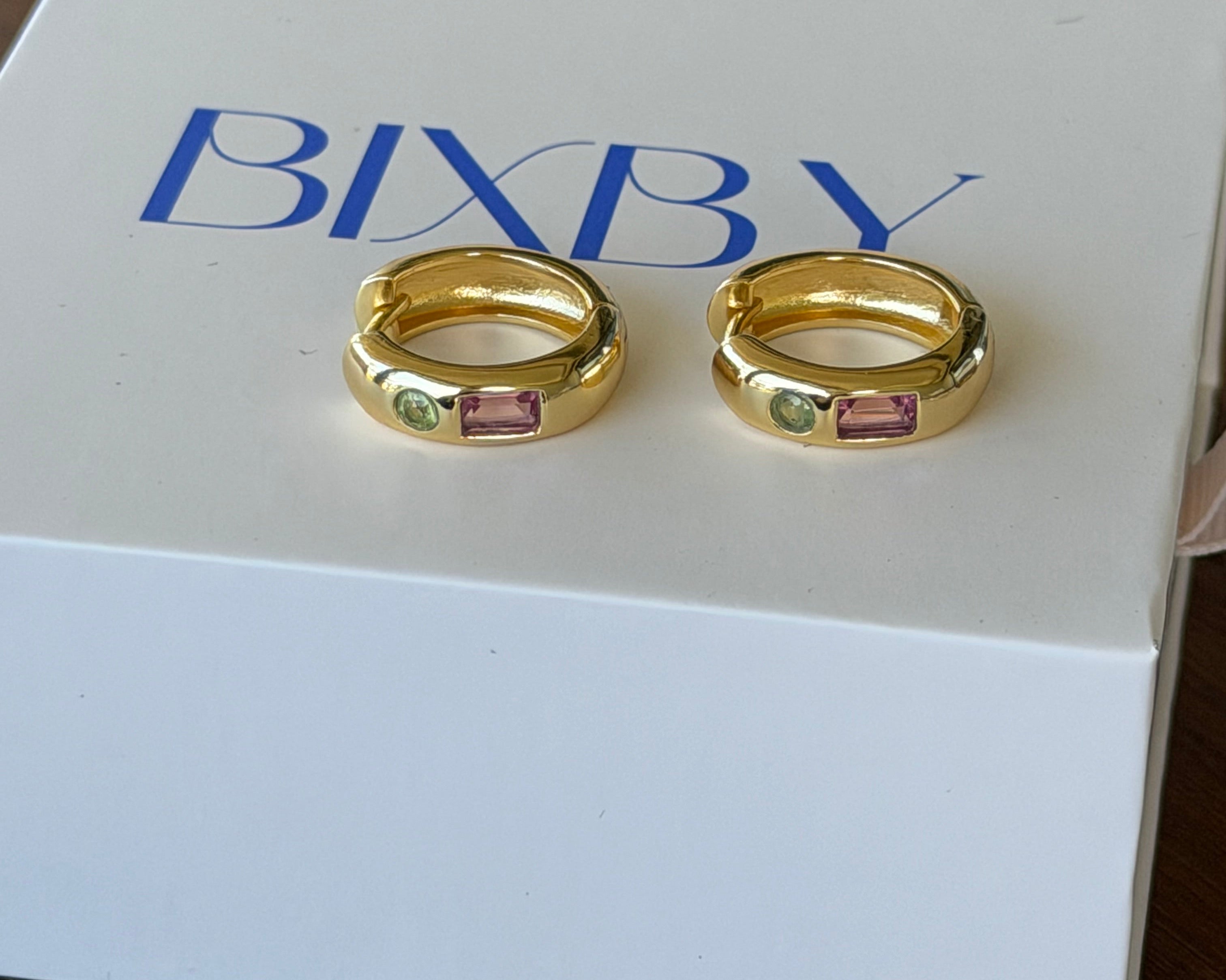 Two gold-toned BIXBY rings with small pink and green gemstones, displayed on a white BIXBY gift box on a dark wooden surface.