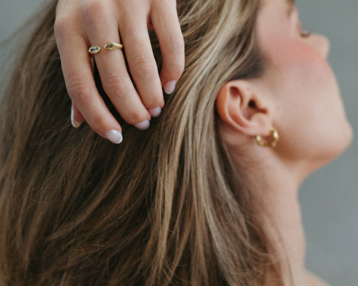 A close-up lifestyle shot of a woman with short brown hair, wearing a delicate gold ring on her finger and a small gold hoop earring, with her hand resting on her head against a soft gray background.