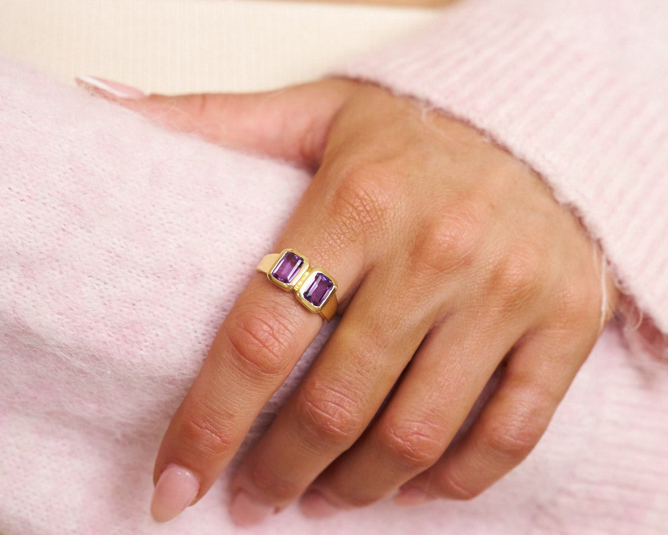 Hand wearing a gold ring with amethyst gemstone on a pink fabric background