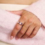 Hand wearing a gold ring with amethyst gemstone on a pink fabric background