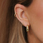 Close-up of a person's ear wearing multiple gold earrings, including a small hoop, a curved ear cuff, and a textured vertical hoop.