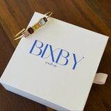 A gold cuff bracelet with colorful rectangular baguette gemstones in pink, red, green, yellow, and blue, resting on a white BIXBY and co. gift box on a dark wooden table.
