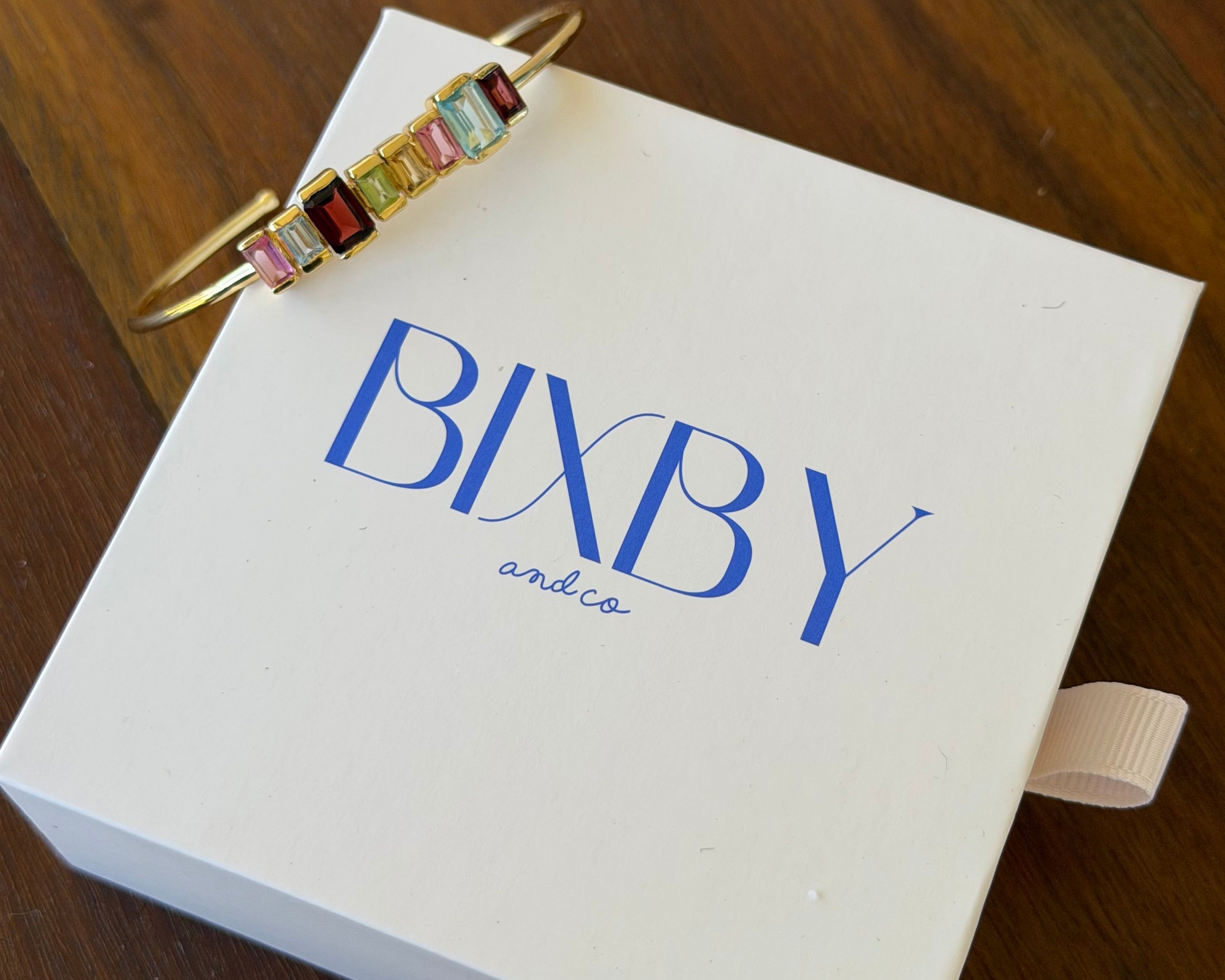 A gold cuff bracelet with colorful rectangular baguette gemstones in pink, red, green, yellow, and blue, resting on a white BIXBY and co. gift box on a dark wooden table.