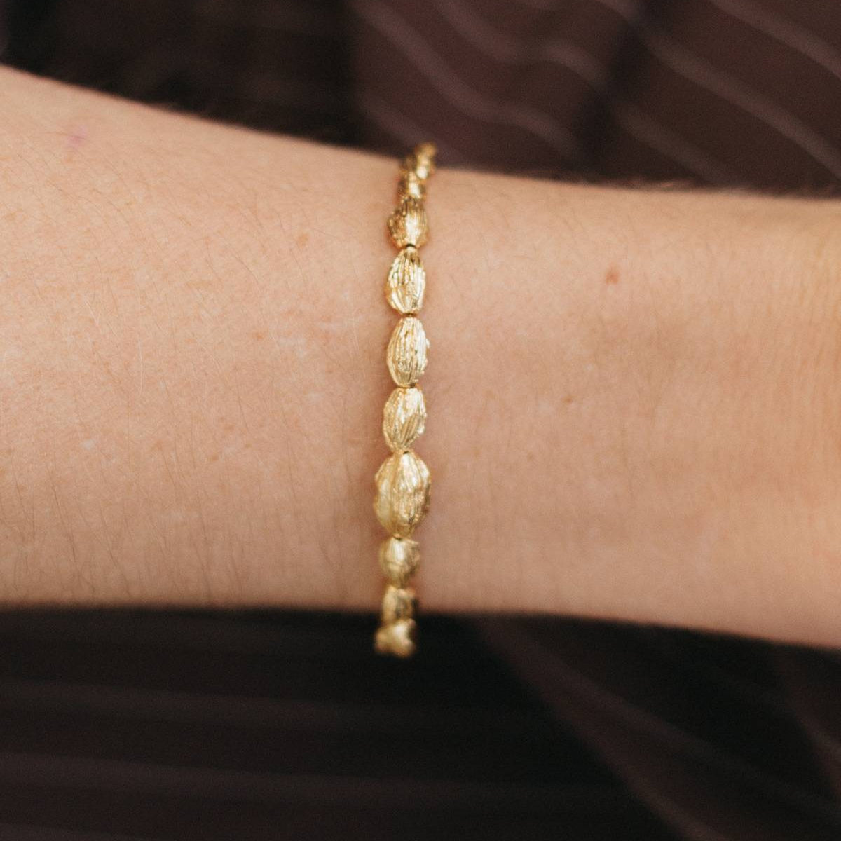A close-up of a person's wrist wearing a delicate gold chain bracelet with textured, organic-shaped links, set against a dark brown striped background.