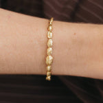A close-up of a person's wrist wearing a delicate gold chain bracelet with textured, organic-shaped links, set against a dark brown striped background.