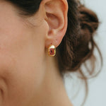 Close-up of a person wearing a gold earring with a red gemstone against a neutral background