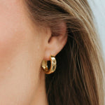 A close-up of a model's ear wearing a pair of polished gold huggie hoop earrings.