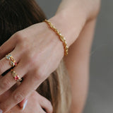 A close-up lifestyle shot of a person's hand and wrist wearing a delicate gold chain bracelet, with matching gold rings on the fingers. The background is a soft, out-of-focus gray.