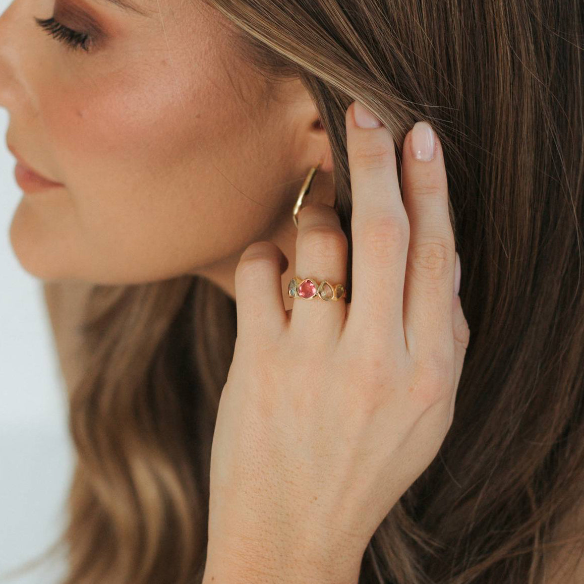 A close-up lifestyle shot of a woman wearing a delicate gold beaded bracelet on her wrist, a gold hoop earring, and a gold ring with small red gemstones, set against her brown hair and tan top.